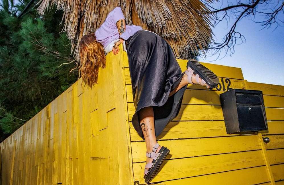 Woman in stylish sandals and lavender hoodie posing on a yellow fence with palm fronds and blue sky in the background in an outdoor setting.
