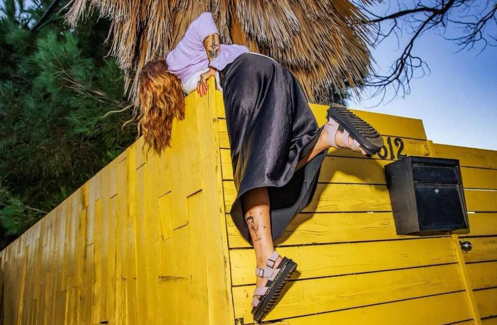 Woman in stylish sandals and lavender hoodie posing on a yellow fence with palm fronds and blue sky in the background in an outdoor setting.
