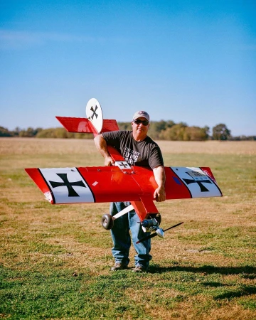 Man holding a red model airplane with black crosses outdoors in a grassy field under a blue sky.