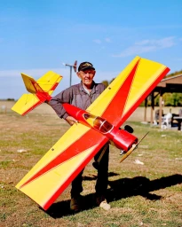 Elderly man wearing a baseball cap holding a large red and yellow model airplane outdoors on a sunny day