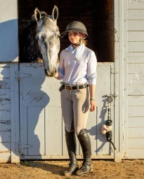 Young equestrian wearing a riding helmet and beige breeches standing beside a grey horse looking out from a stable stall.