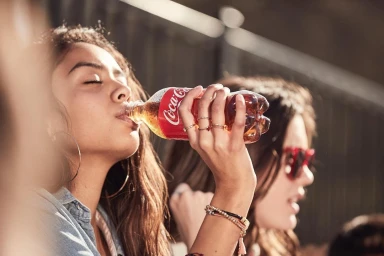 Young woman drinking from a Coca-Cola plastic bottle outdoors with friends nearby in bright natural sunlight.