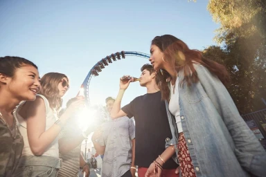 Low-angle shot of a group of friends holding glass bottles at an amusement park with a roller coaster under a clear sky.