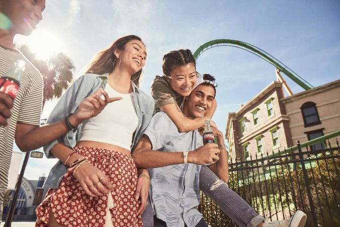 Four diverse young friends smiling and laughing together outdoors, holding glass bottles with a roller coaster track visible in the background.
