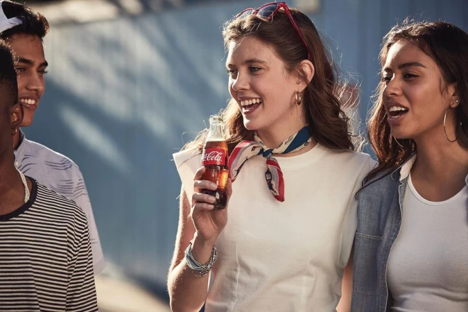 Smiling young woman holding a glass Coca-Cola bottle surrounded by friends outdoors in New York