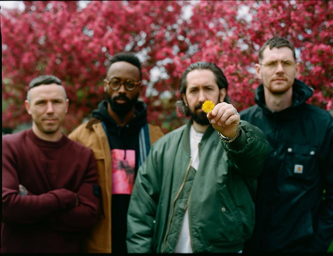Four adult males pose outdoors in front of a tree with red blossoms. One man holds a yellow dandelion in his hand.