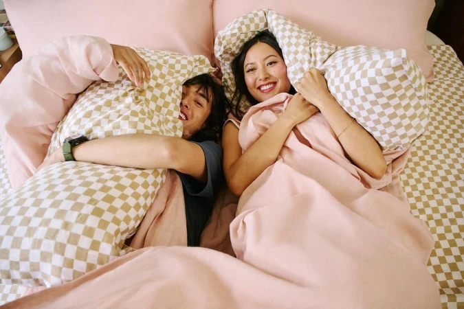 Man and woman smiling in bed holding checkered pillows wrapped in pink bedding