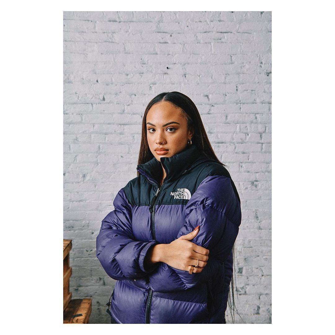 Portrait of a young adult woman in a stylish purple puffer jacket with a black collar against a white brick wall in a studio setting.