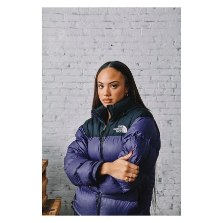 Portrait of a young adult woman in a stylish purple puffer jacket with a black collar against a white brick wall in a studio setting.