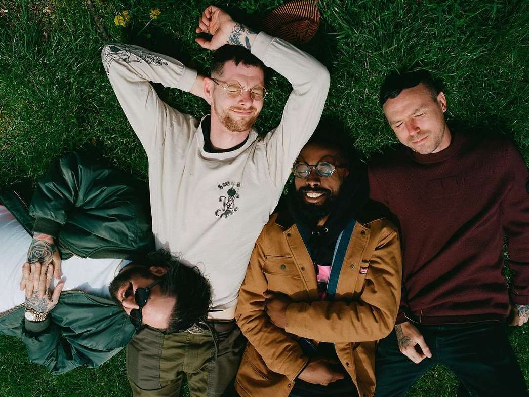 Four adults pose in the grass together outdoors. Men wear casual clothing and fashionable eyewear in a lifestyle setting with bright, natural light.