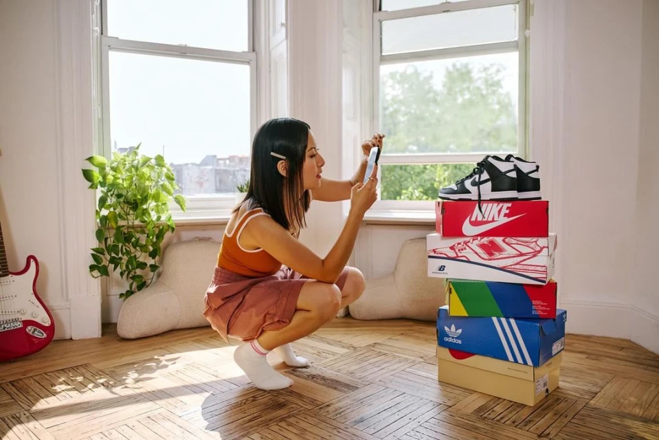 Woman squats indoors taking a smartphone photo of stacked branded shoe boxes with a pair of Nike Dunk sneakers on top near a window.