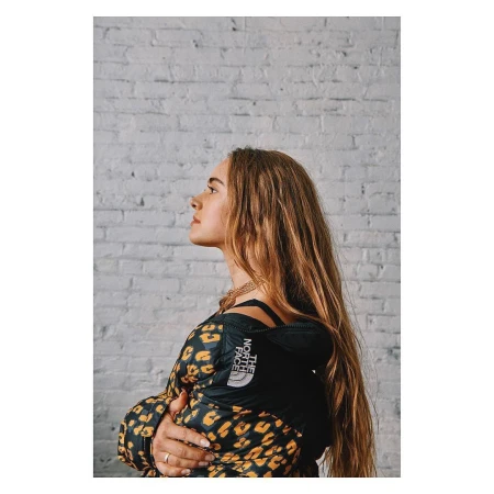 Profile portrait of a young woman wearing a leopard print puffer jacket against a painted white brick wall.