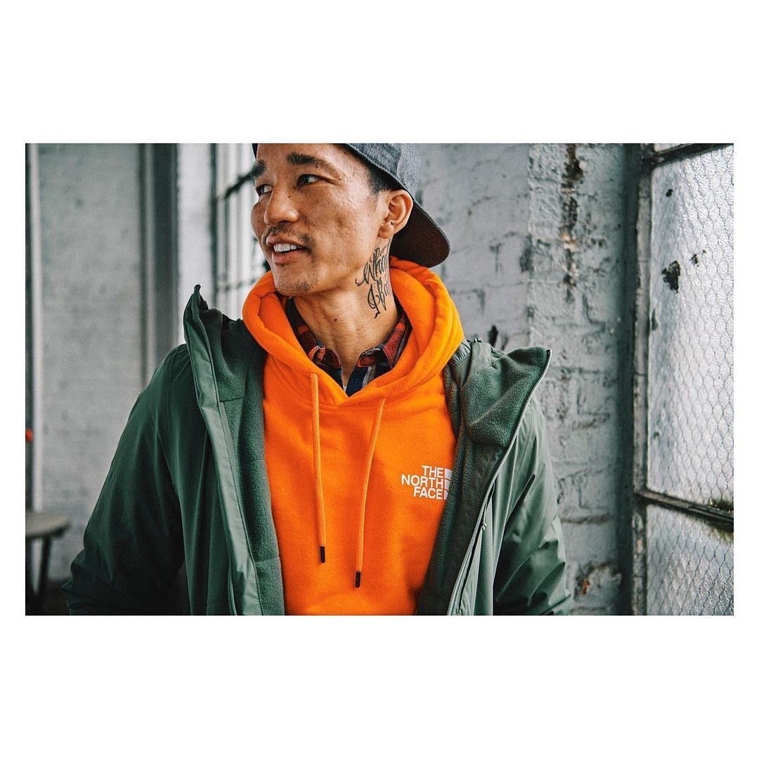Young Asian man in a bright orange hoodie and green jacket stands near a window with a brick wall in the background looking away.