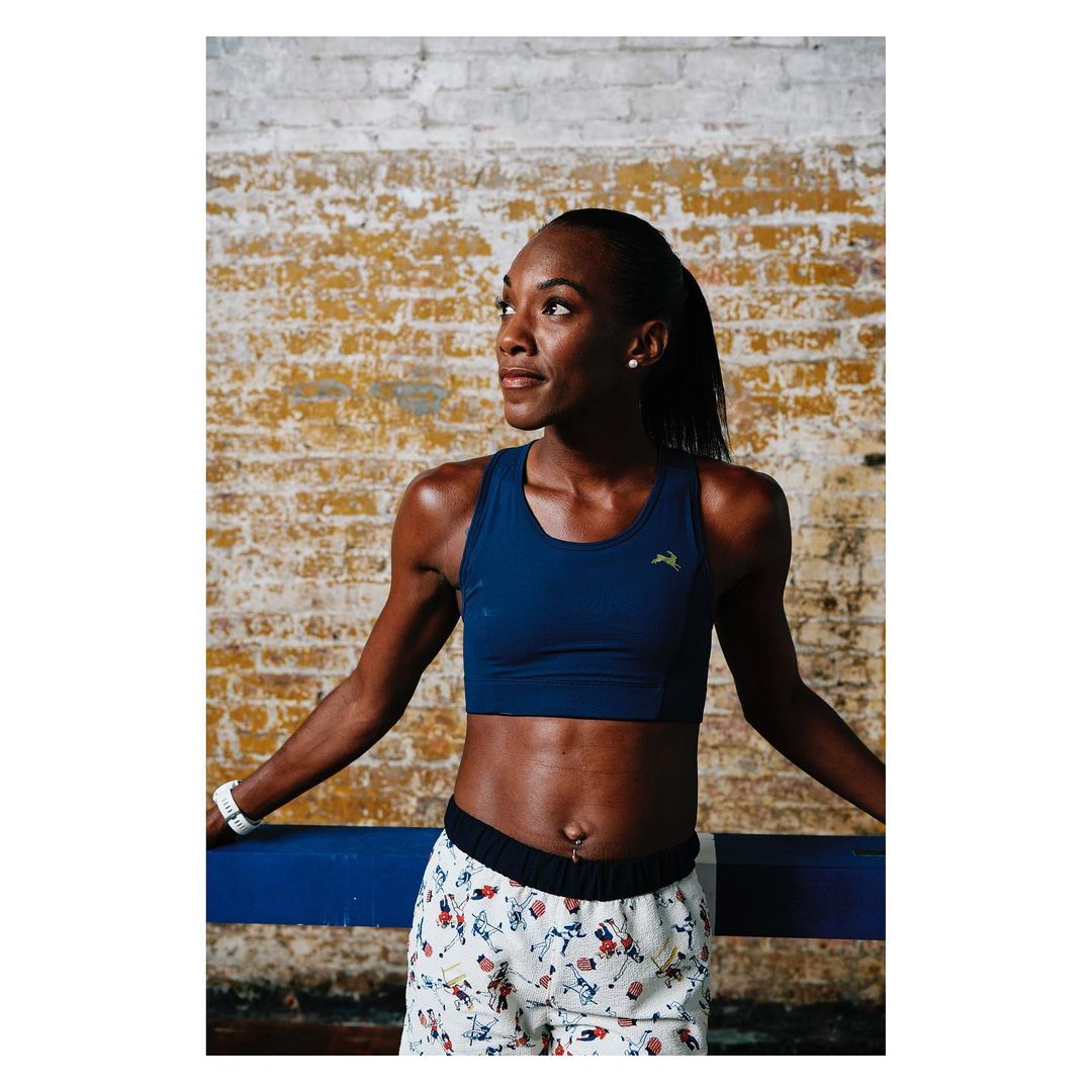 A fit African-American woman in a crop top and patterned shorts leans against a blue bar indoors, with a brick wall behind her.
