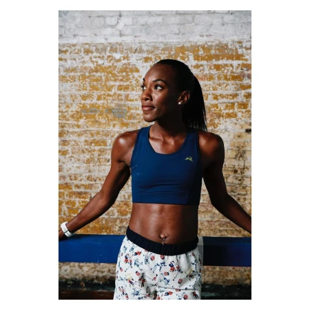 A fit African-American woman in a crop top and patterned shorts leans against a blue bar indoors, with a brick wall behind her.