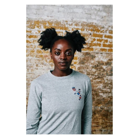 Portrait of a black woman with pigtails wearing a grey long-sleeve t-shirt against a textured brick wall