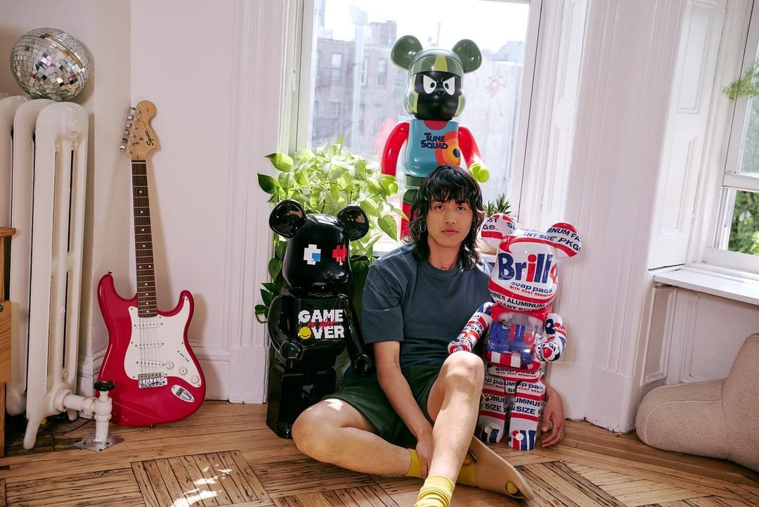 Young adult poses indoors with three toy figures: one black bear-shaped, one white bear-shaped, and one Space Jam character figure.