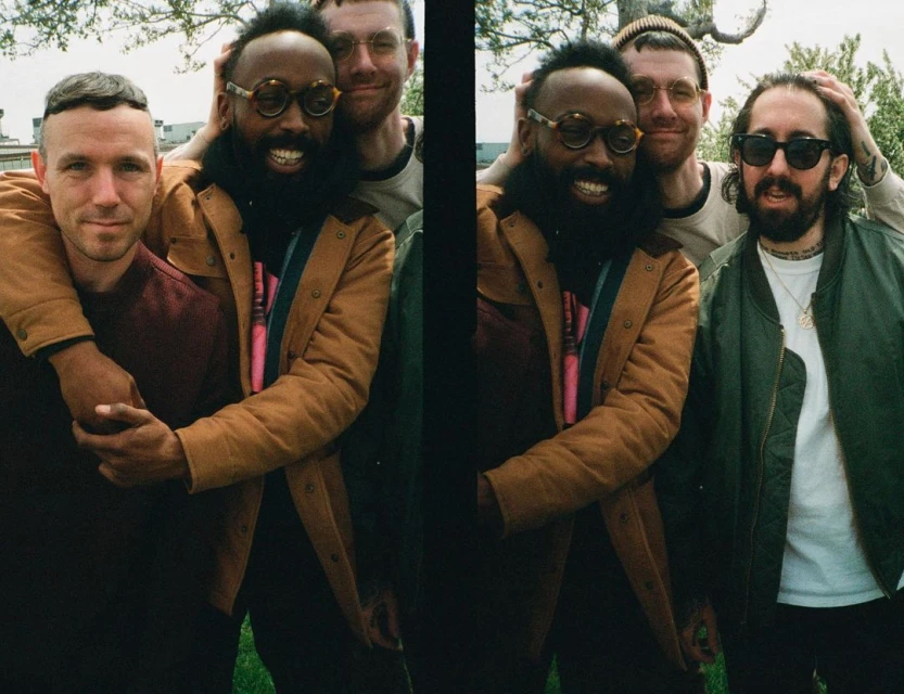 Four adult men pose outdoors with a brick building and trees in the background; three of the men have arms around each other.