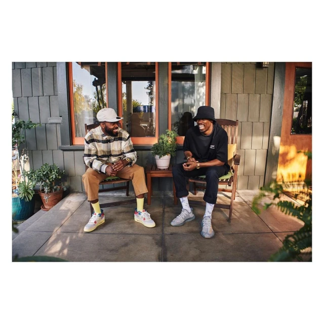 Two adult males are sitting on wooden chairs outdoors on a porch, with potted plants, enjoying a conversation in daytime lighting.