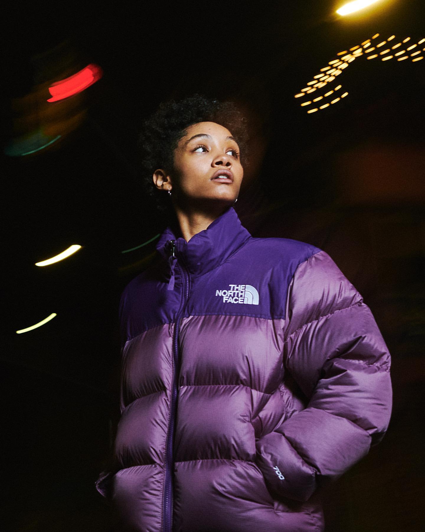 A woman with dark, short hair poses in a two-tone purple The North Face puffer jacket against a blurry, dark outdoor backdrop at night.