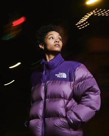 Woman wearing a purple puffer jacket against a dark night background with blurred lights by Ben Rayner.