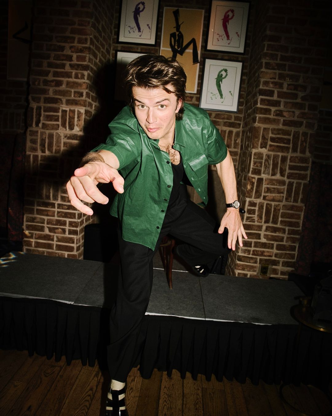 Full-length shot of a man in green button-down and black pants, pointing. Brick wall and framed art in the background.