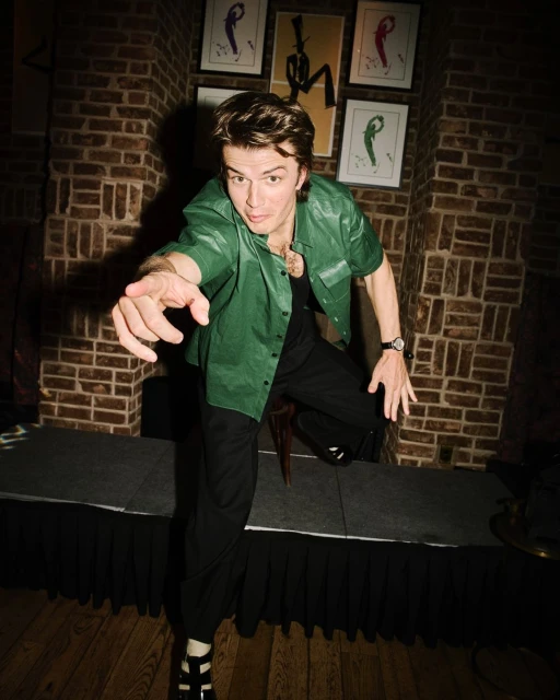 Full-length shot of a man in green button-down and black pants, pointing. Brick wall and framed art in the background.