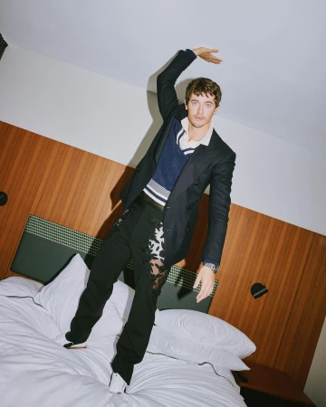 A man in a dark double-breasted blazer and patterned trousers standing on a hotel bed, holding his hand against the ceiling.
