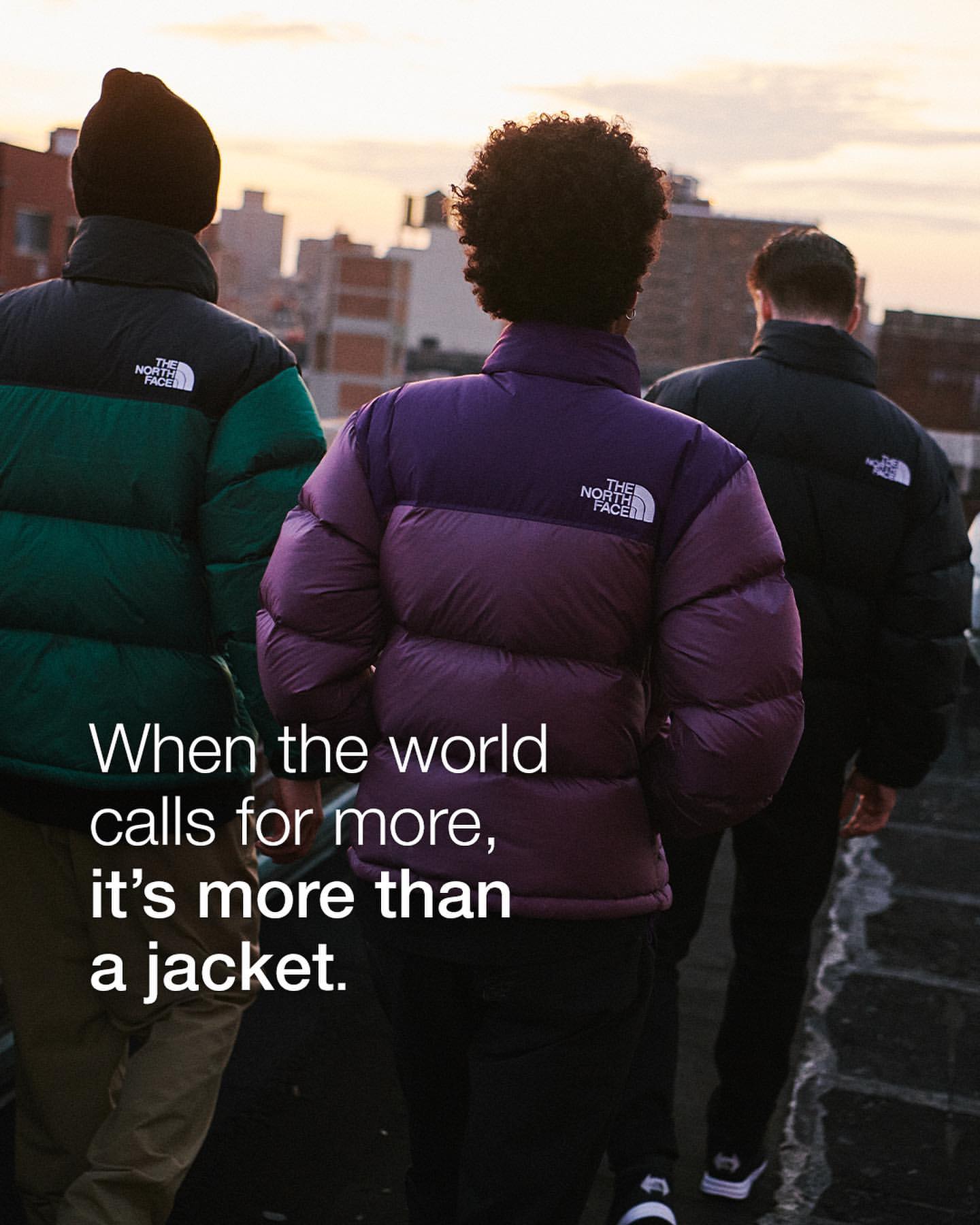 Three people wearing The North Face down puffer jackets stand on a rooftop with buildings and a sunset in the background.