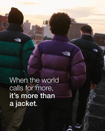 Three people wearing The North Face down puffer jackets stand on a rooftop with buildings and a sunset in the background.