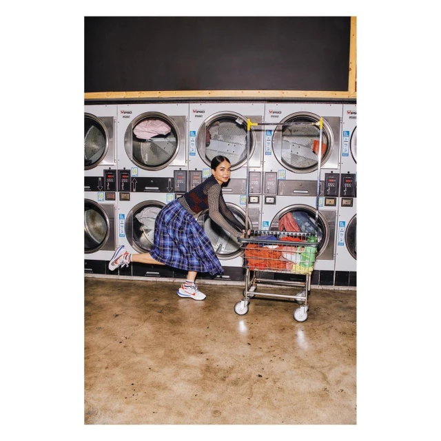 Woman in a blue plaid skirt playfully pushes a laundry cart through a laundromat, surrounded by rows of stainless steel washers.