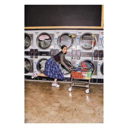 Woman in a blue plaid skirt playfully pushes a laundry cart through a laundromat, surrounded by rows of stainless steel washers.