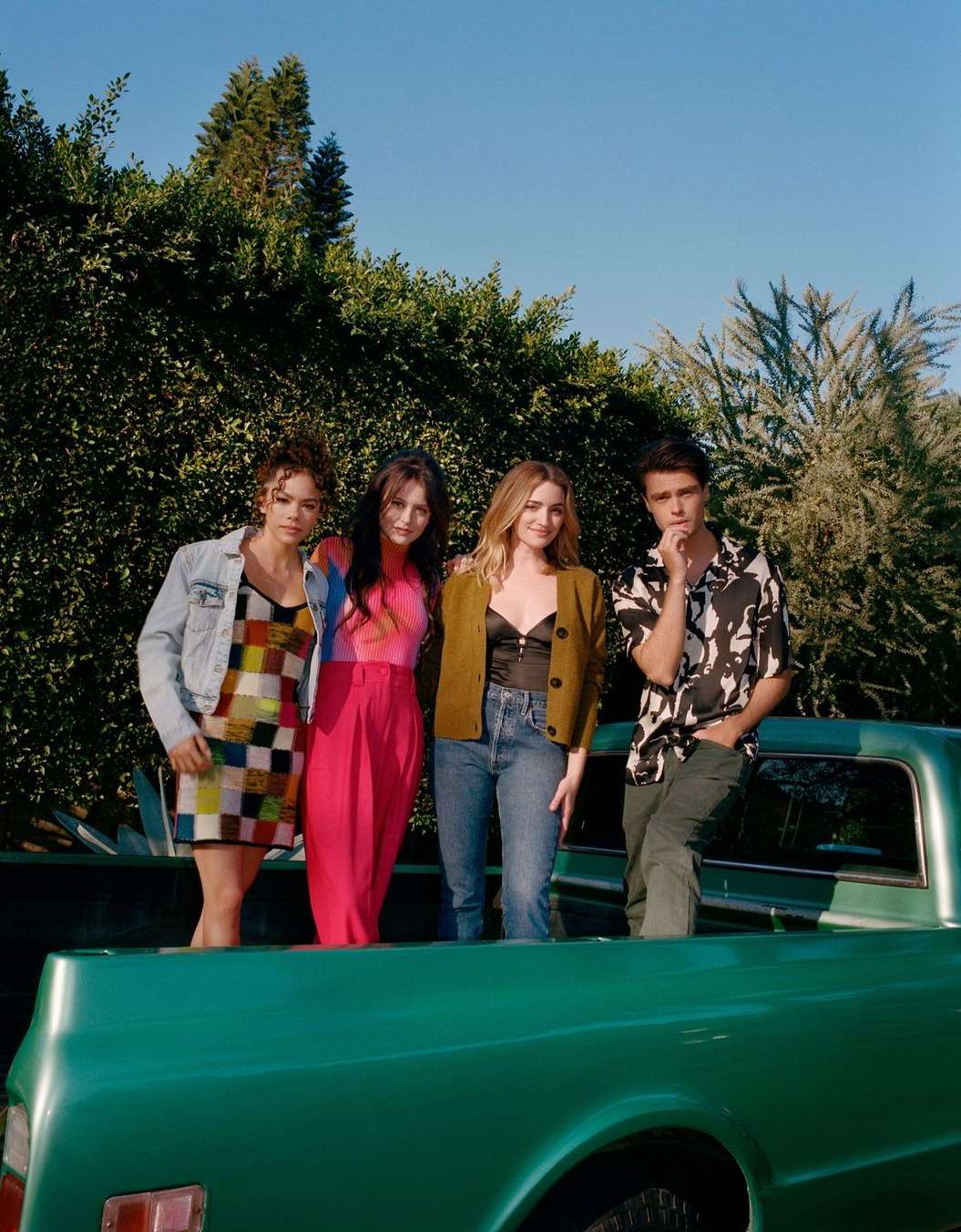 Group of four young adult friends stand in the bed of a vintage green pickup truck against a backdrop of green trees and a blue sky.