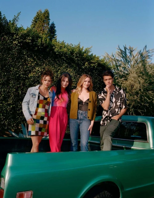 Group of four young adult friends stand in the bed of a vintage green pickup truck against a backdrop of green trees and a blue sky.