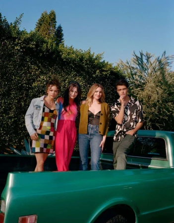 Group of four young adult friends stand in the bed of a vintage green pickup truck against a backdrop of green trees and a blue sky.