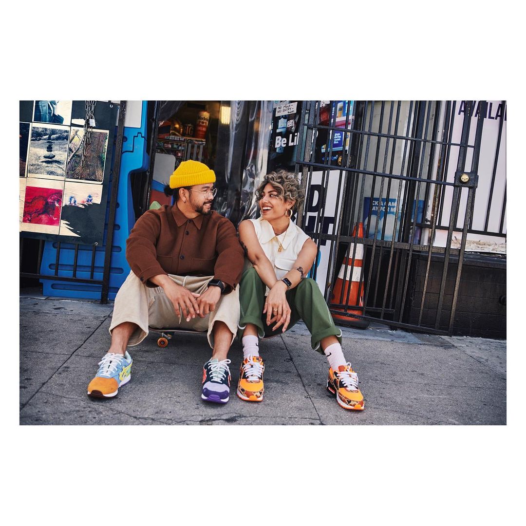 Two people sit outdoors on a city street smiling at each other, with visible shoes, clothing, and an urban environment in the background.