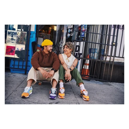 Two people sit outdoors on a city street smiling at each other, with visible shoes, clothing, and an urban environment in the background.