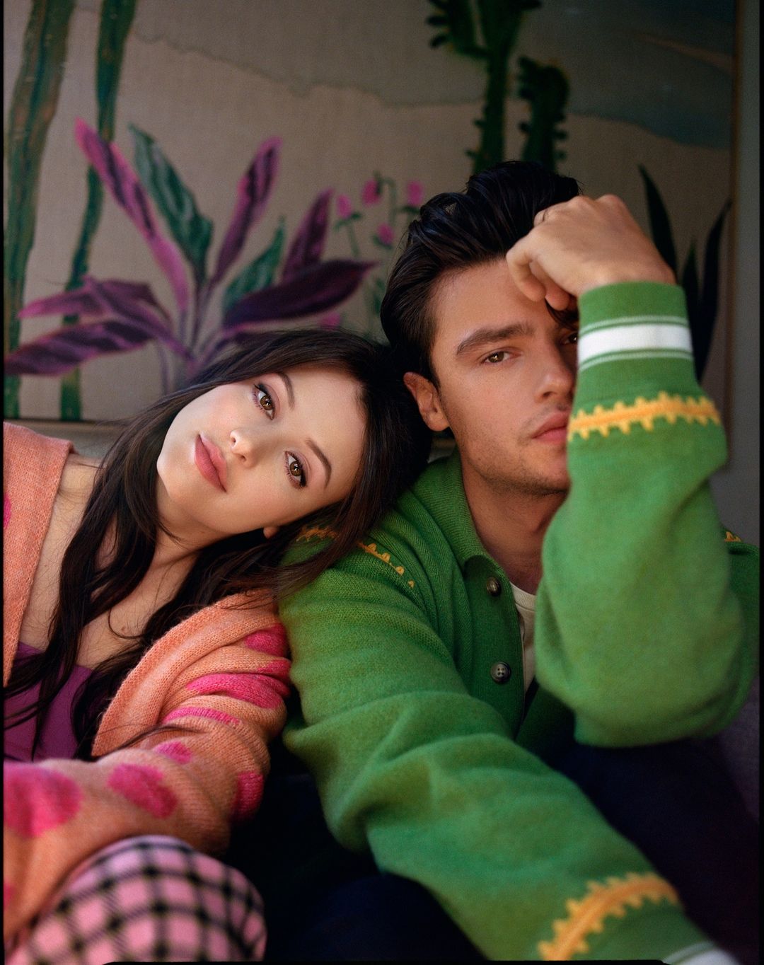 Portrait of a young couple seated closely together, facing the camera, showcasing a colorful floral backdrop and stylish vintage attire.