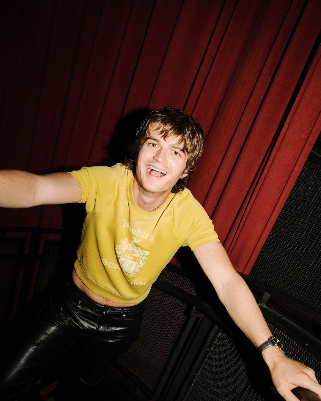 Smiling man in a yellow shirt and black pants poses against a red curtain with a metal railing. The lighting highlights his expression.