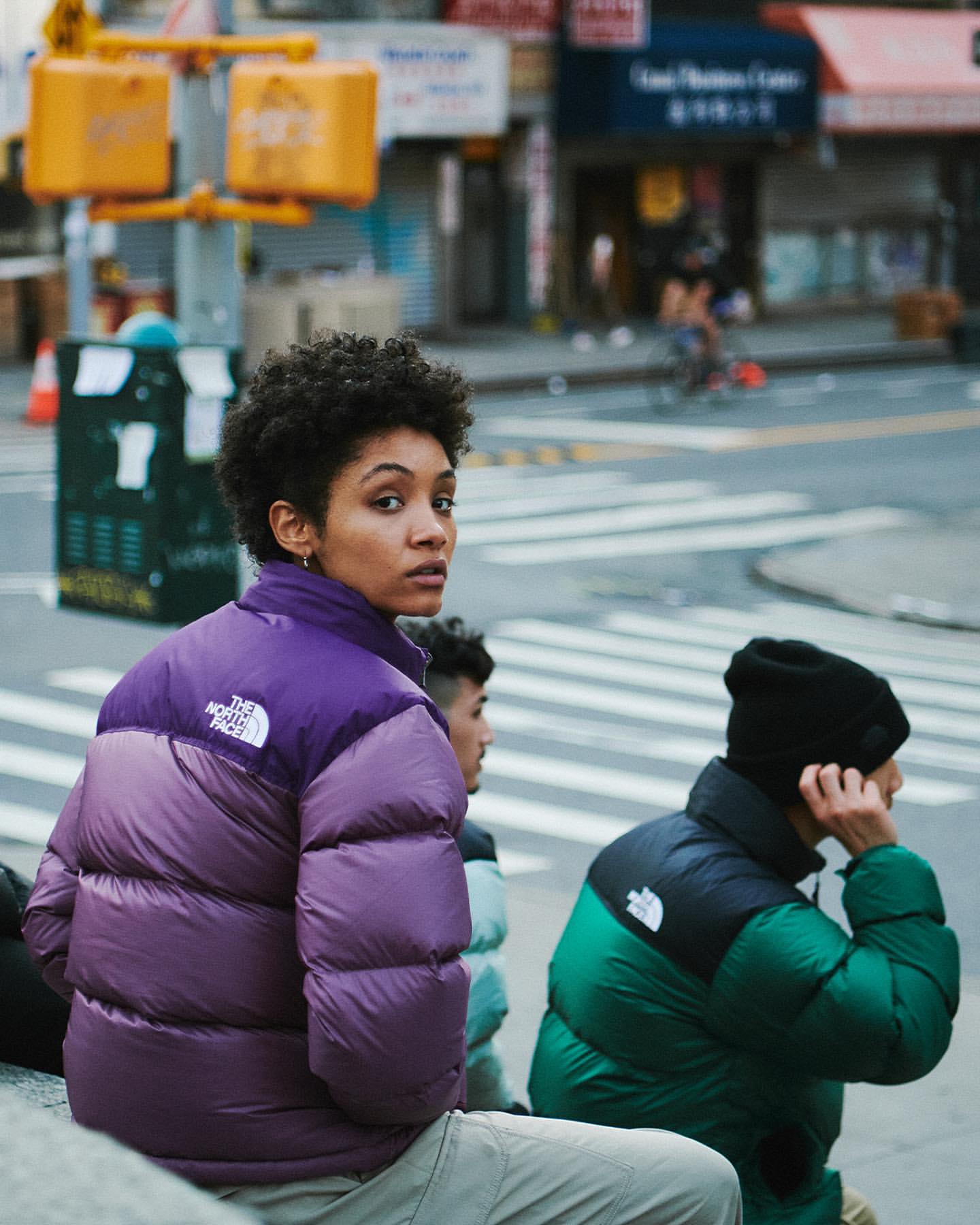 Outdoor shot of a young adult woman in a purple puffer jacket, looking over her shoulder, with people and urban street background.