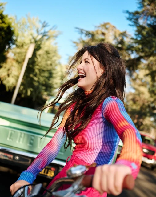 Woman riding bicycle on tree-lined street with vintage truck. She has long brown hair and wears a colorful striped shirt and pink bottoms.