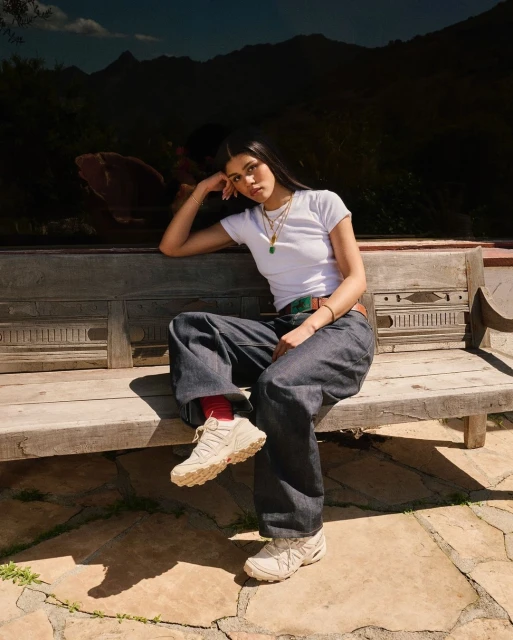 A young woman sits on a wooden bench outdoors wearing a white t-shirt, baggy jeans, and sneakers, with mountains in the background.