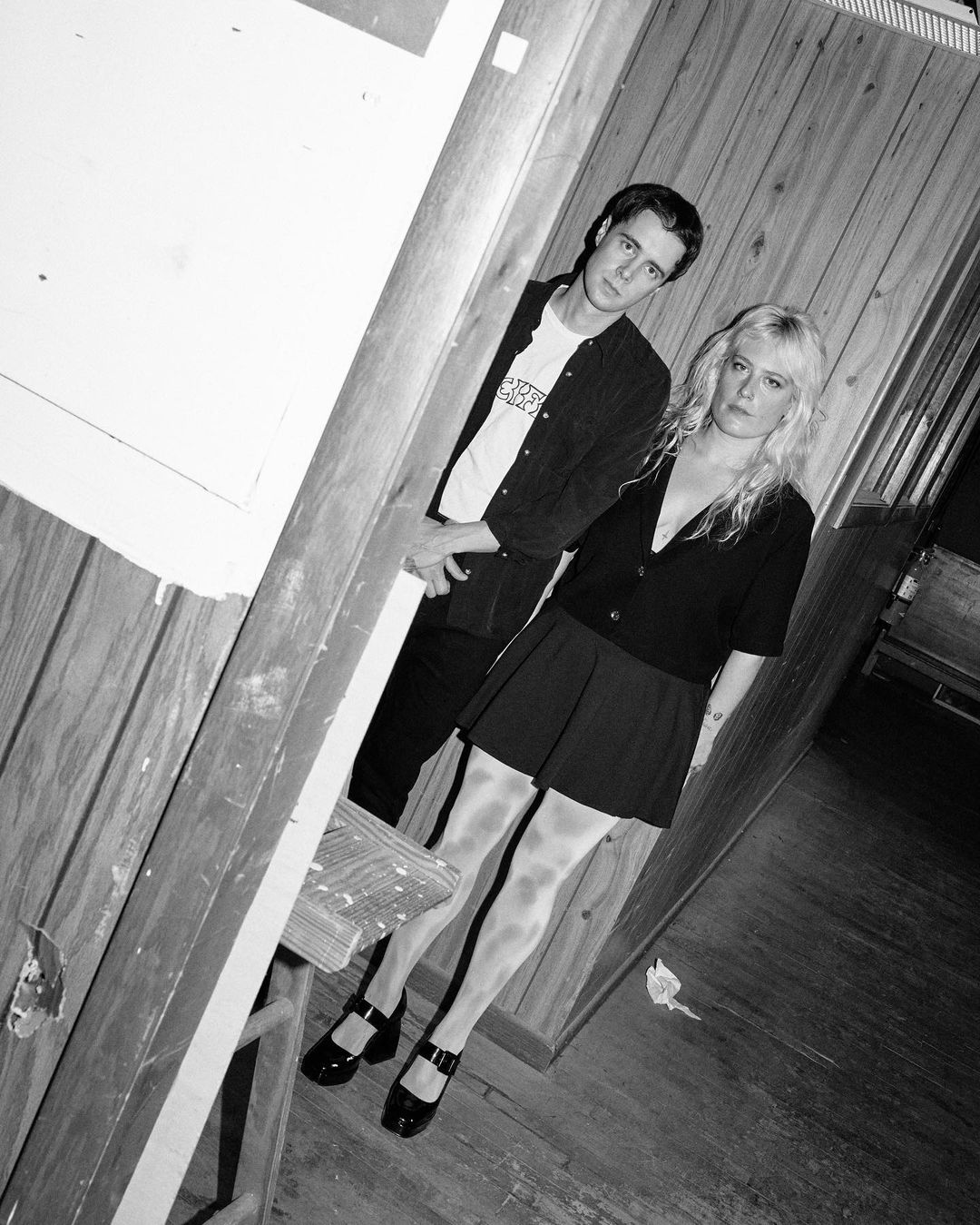 Monochrome portrait of a young couple standing together indoors in a wood-paneled room, glancing toward the camera in a studio shot.