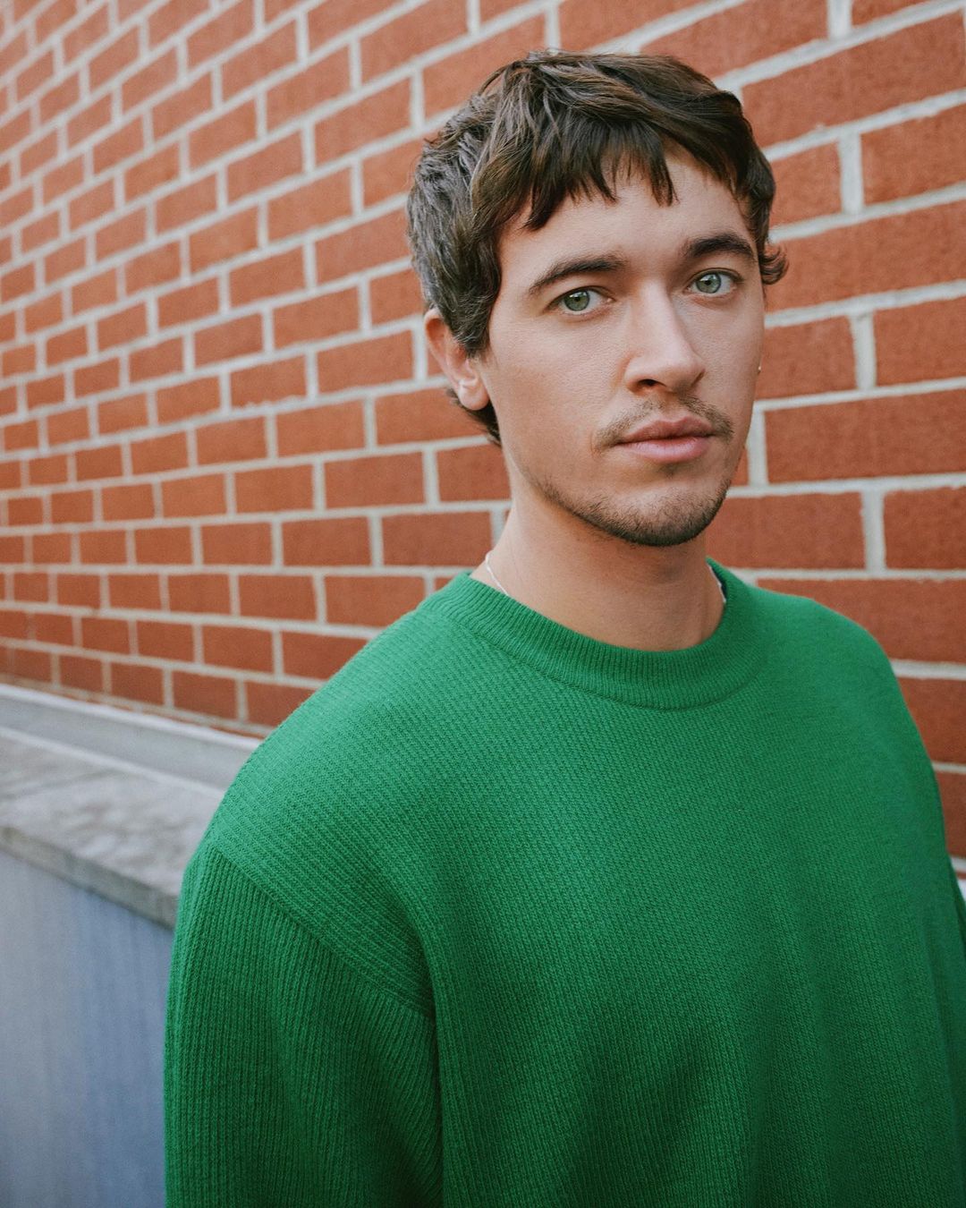 Headshot of a young adult male with green eyes and brown hair, wearing a green sweater, posing against a brick wall outdoors.