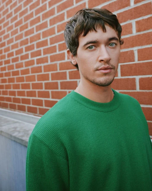 Headshot of a young adult male with green eyes and brown hair, wearing a green sweater, posing against a brick wall outdoors.
