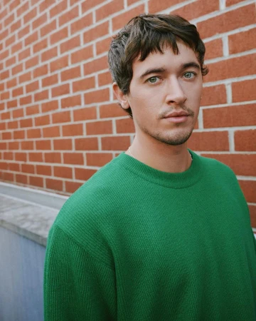 Headshot of a young adult male with green eyes and brown hair, wearing a green sweater, posing against a brick wall outdoors.