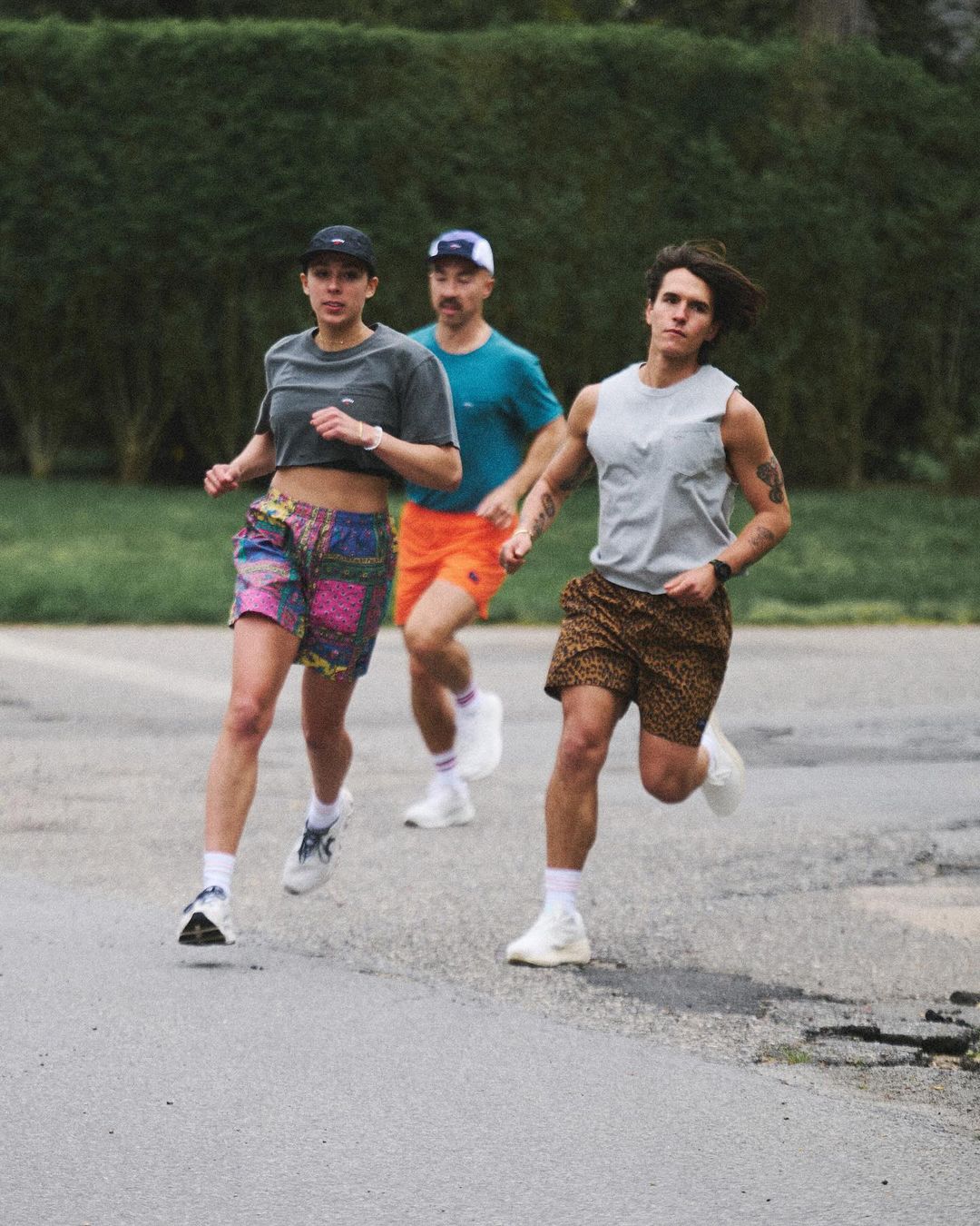 Three people running outdoors on a paved surface with green foliage in the background.  Runners are wearing athletic clothing and sneakers.