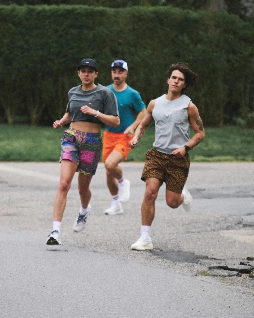 Three people running outdoors on a paved surface with green foliage in the background.  Runners are wearing athletic clothing and sneakers.