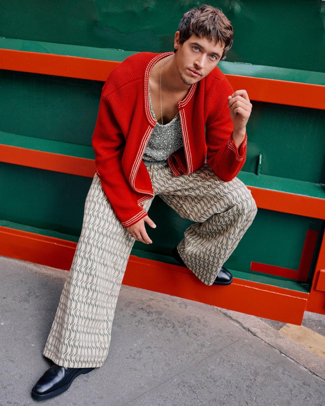 A fashionable young man with dark hair and blue eyes is posed indoors wearing a red cardigan, patterned pants, and black shoes.
