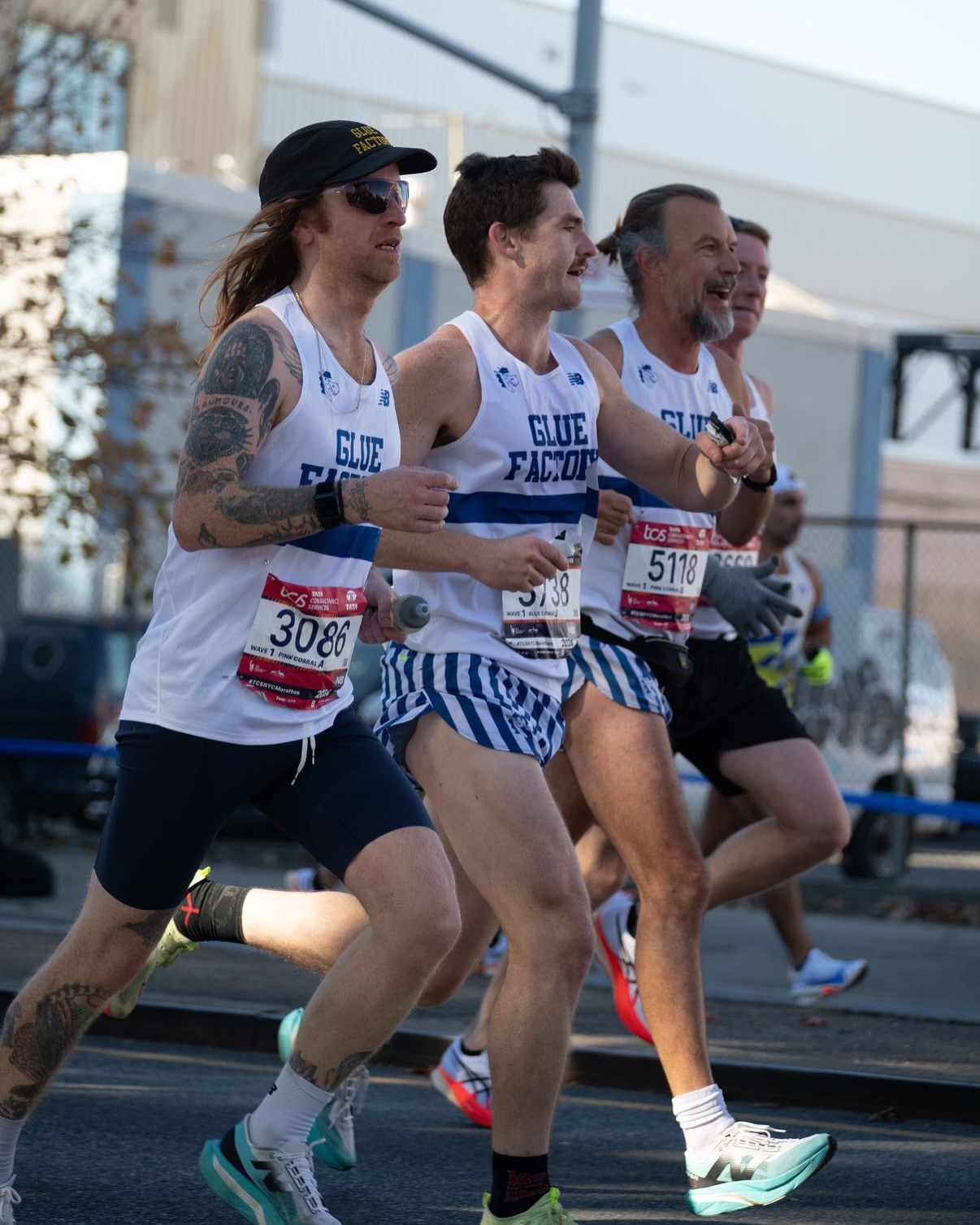 A group of male runners in athletic wear compete in an outdoor road race, wearing numbered bibs and running shoes on a sunny day.